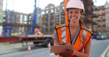 Kasketli ve güvenlik yeleği giyen kadın inşaat işçisinin portresi panoya yazılıydı. Sokakta kameraya gülümseyen büyük yerleşim yerinin yanında duruyordu. 4k