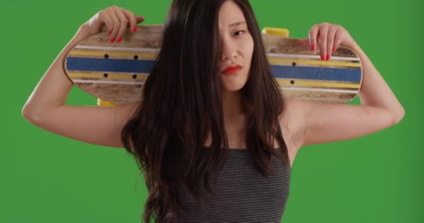 肩にスケートボードを担いで緑の画面上のカメラを見ている若いアジアの女性のいくつか スケートボードとクールな中国の女性は緑の画面に隔離された — ストック動画