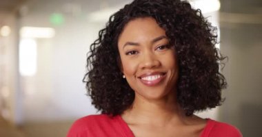 Kameraya gülümseyen kıvırcık saçlı, sevimli siyah bir kadın. Kapalı mekan ortamında gülümseyen Afrikalı genç bir kadının yakın çekimi. 4k