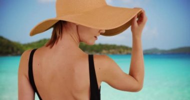 Virgin Adaları, St John 'da güzel bir plajda duran sarkık şapkalı bin yıllık kadının dikiz görüntüsü. Tropikal okyanusa bakan bir turist. 20 'li yaşlarda, güneş şapkalı ve siyah mayosu olan bir kadın. 4k
