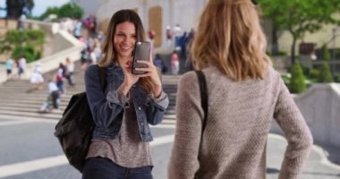 Genç beyaz bir kadın İtalya, Roma 'daki arkadaşının fotoğrafını çekmek için kameralı telefon kullanıyor. Muhteşem esmer, İtalya, Roma 'da seyahat ederken sarışın arkadaşının fotoğrafını çekiyor. 4k