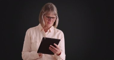 Gri arka planda internette gezinmek için tablet kullanan çekici bir yaşlı kadın. Koyu gri arka planda kablosuz ped kullanan nazik olgun bir bayan. 4k