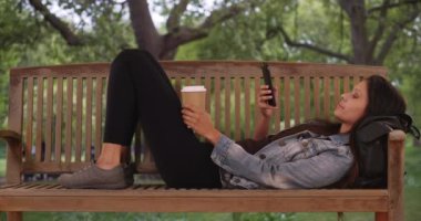 Kot ceketli beyaz kadın parktaki bankta uzanıyor ve sosyal medyada geziniyor. Kadın yastık niyetine sırt çantasıyla uzanırken kahveyi tutuyor. 4k