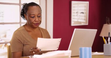 Yaşlı siyah bir kadın vergilerini ödemek için telefonunu ve laptopunu kullanıyor. Yaşlı bir Afrikalı Amerikalı kadın, muhasebe yapmak için bilgisayarını ve cep telefonunu kullanıyor. 4k