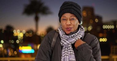 Geceleri şehir merkezinde sıcak kıyafetler giyen yaşlı siyah bir kadın. Geceleri soğuk havada yaşlı bir Afrikalı Amerikalı kadının portresi. 4k