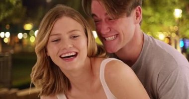 Parkta arkadan gülüşen erkek sevgiliye yakından bakın. Geceleri dışarıda kucaklaşan, gülümseyen ve gülen genç bir çiftin portresi. 4k