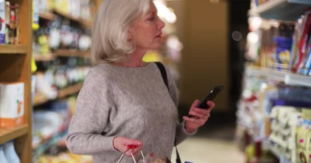 Старша Жінка Дивиться Продуктовий Прохід Мобільним Телефоном Руці Літня Жінка — стокове відео