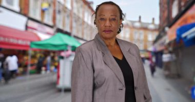 Brixton Caddesi 'ndeki markette kameraya gülümseyen olgun bir iş kadını. Takım elbiseli küçük işletme sahibi açık havada portre için poz veriyor. 4k