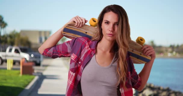 海によって屋外スケートボードでポーズ白い千年紀の少女の肖像画 湾で肩の上にスケートボードを保持する流行の女性スケーターの肖像画 — ストック動画