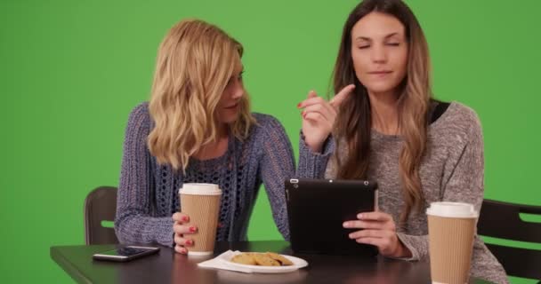 一对快乐的女孩坐在那里喝酒 看着平板电脑准备好了彩色按键作曲 两个漂亮的高加索女人使用便携式平板电脑 并在绿色屏幕上的桌上社交 — 图库视频影像