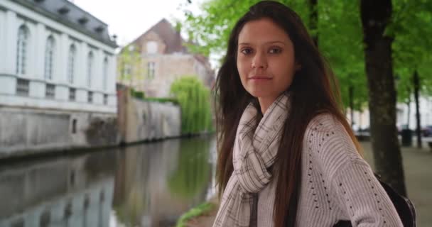 ベルギーのブルージュの運河によるグレーのセーターの美しい女性は目の接触を作ります 街の通りに彼女のバックパックを持つ女性旅行者の肖像画 — ストック動画