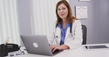 Bilgisayarda yazı yazan ve kameraya gülümseyen güzel bir kıdemli doktorun portresi. Ofiste oturan ve kameraya bakan çekici tıp hekimlerine yakın çekim.