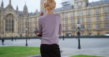 Londra caddesinde genç ve çekici bir iş kadınının dikiz görüntüsü. Telefonuyla Westminster Palace 'a bakan bir kadın yönetici. 4k