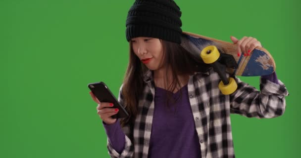 携帯電話を使用している間 肩の上にスケートボードを運ぶ若いアジアのヒップスターは 画面上のテキストメッセージを読み取ります 緑の画面上でスマートフォンを使用している間 スケートボードを保持しているリラックスした中国の女の子 — ストック動画