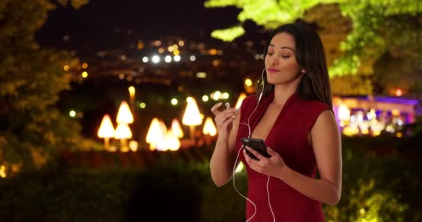 夜の外で音楽を聴く魅力的な笑顔の若いラティーナの女性 スマートフォンで音楽を聴くイヤホン付きの明るい女性の肖像 — ストック動画