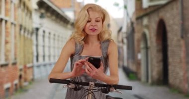 Belçika 'nın Bruges kentindeki eski caddelerde yol tarifi için akıllı telefon kullanan turist bir kadın bisikletiyle dolaşıyor. Cep telefonunda elbise konsültasyonu haritasında güzel bir kadın. 4k