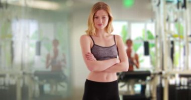 Spor salonunda kollarını kavuşturup kameraya bakan atletik giysili genç, beyaz bir kadın. Vücut geliştirme merkezinde çalışan sarışın bir kadın. 4k