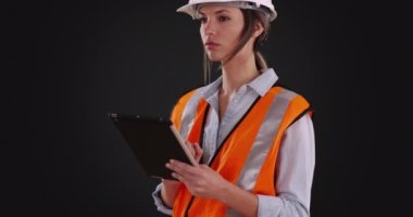 Turuncu yelekli ve kasklı bir kadın gri arka planda tablet üzerinde çalışıyor. Koyu gri arka planda çalışan kadın inşaat işçisi. 4k