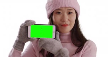 Kışın giyinmiş gülümseyen Asyalı kadın elinde beyaz arka planda yeşil ekranlı akıllı telefon tutuyor. Eldiven ve süveter giyen mutlu bir milenyum cep telefonuyla ekranı fotokopi alanıyla paylaşır. 4k