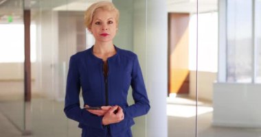 Ofis binasında cam bölmenin yanında duran kadın yönetici gülümseyen kameraya bakıyor. Modern ofis koridorunda cep telefonu pozu veren çekici bir iş kadını. 4k