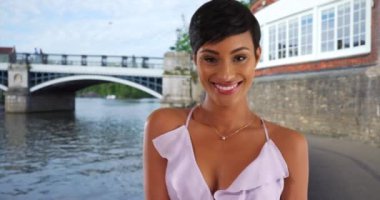 Windsor, İngiltere 'de güzel gülümseyen siyah kadın yaz elbisesi içinde poz veriyor. Thames nehrinin kenarında kameraya gülümseyen güzel Afrikalı kadın portresi. 4k