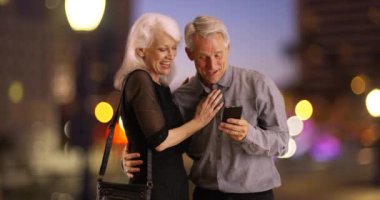 Gece vakti akıllı telefon kullanan mutlu yaşlılar. Yaşlı bir çift gece şehir dışına çıkıyor ve cep telefonu kullanıyor. 4k