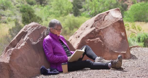 一个健康的老年妇女在峡谷里有时间在户外看书的画像 快乐成熟的女人 坐在大自然的岩石旁 享受着书本和咖啡 — 图库视频影像