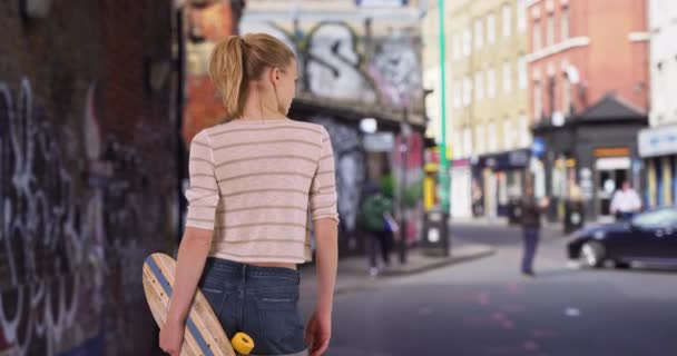スケートボードを保持する彼女の20代の若いヒップスター女性のリアビュー スケートボードでロンドンの通りに立つ白人女性 — ストック動画