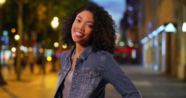 穿着牛仔夹克的快乐的非洲裔美国女人在夜晚站在城市街道上对着镜头微笑 美丽的年轻千年的笑和微笑在香蕉丽舍大街上的夜晚 — 图库视频影像