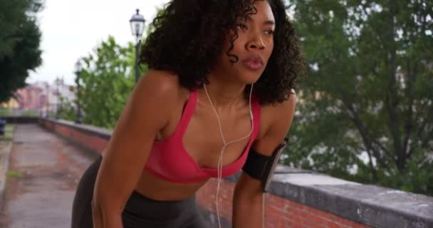 非洲裔美国妇女慢跑者在城市里跑步时停下来检查健身跟踪器 跑步者用智能手机和耳机屏住呼吸 然后跑出屏幕 — 图库视频影像