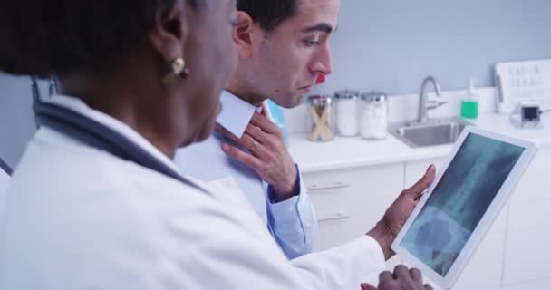 脊髄と骨盤の患者X線を表示するために電子タブレットを使用して中高年の黒人医師 若いですLatino患者見ますX線の彼の体上の電子ノートブックタブレット — ストック動画