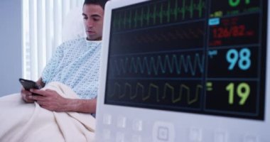 EKG monitörüne bağlıyken genç Latin bir erkeğin cep telefonuyla mesajlaşmasını yakından izleyin. Hastanenin monitöründeki EKG değerlerinin portresi.