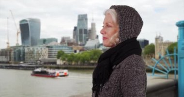 Kış tatilinde Londra 'da Thames nehrinin üzerinden bakan yaşlı bir kadın. Soğuk İngiliz tatilinde ufuk çizgisine hayran olan olgun bir turist. 4k