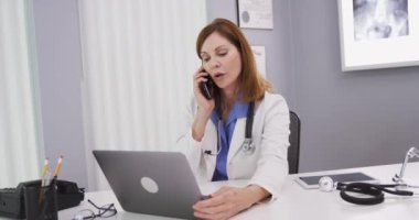 Meslektaşınla akıllı telefondan konuşurken latop bilgisayar kullanan çekici orta yaşlı bir doktor. Ofisinde oturan yakışıklı kıdemli doktor defter bilgisayarına bakıyor ve cep telefonuyla sohbet ediyor.
