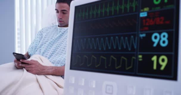 Ekgモニターにフックアップしながら 携帯電話上の若い男性ラテン語患者のテキストメッセージの近いビュー 病院のモニターで読むEcgバイタルの肖像 — ストック動画