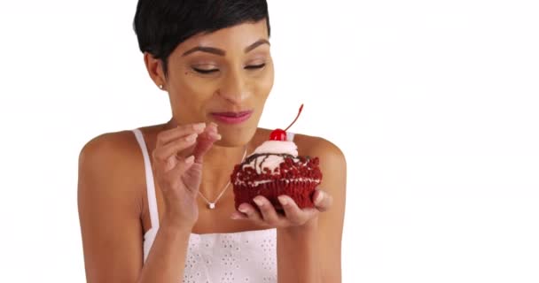 魅力的な黒の女性がお食事を楽しんで閉じます コピースペースとスタジオでカップケーキの霜を試飲 20代の女性の肖像画は 大きな赤いベルベットのカップケーキを保存し マラッシーノの桜を吸います — ストック動画