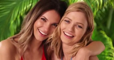 Sarışın arkadaşının kollarında, tropikal ortamda kameraya gülümseyen çekici esmer kız. Bir çift kadın en iyi arkadaş palmiye ağaçlarının yanında mutlu poz veriyorlar. 4k