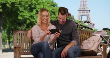 Paris 'teki neşeli genç çift akıllı telefondan komik videolar izliyor. Beyaz erkek ve kadın dışarıda dinleniyor ve teknolojiyi kullanıyor. 4k
