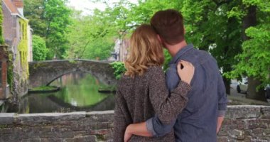 Genç evli çiftin dışarıda gülümseyerek rahatlayışının arka görüntüsü. Neşeli beyaz erkek ve kadın Bruges 'deki kanala bakıyorlar. 4k