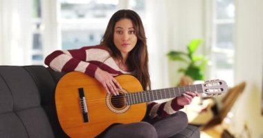 Kanepede oturan, kucağında akustik gitar tutan genç Latin kadının portresi. Oturma odasında uzanan, enstrümanı tutan ve kameraya bakan hoş bir kadın sanatçı. 4k
