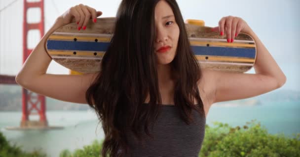 ゴールデンゲートブリッジで頭と肩の後ろにスケートボードでポーズグラニースケーターの女の子 クールな千年アジアの女性は カリフォルニア州サンフランシスコでカメラのためのスケートボードを保持 — ストック動画