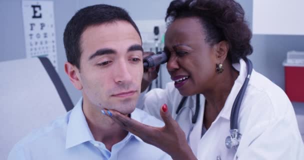 若い患者の耳を検査するために聴診器を使用してアフリカの高齢医師の肖像画 定期検査中に耳をチェックされた魅力的なラテン系患者の閉鎖 — ストック動画