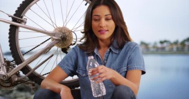 Okyanus kıyısında bisikletiyle oturan çekici İspanyol kadın kameraya bakıyor. Gündüz vakti su şişesi tutan Latin bir kadın bisikletin yanında dinleniyor. 4k