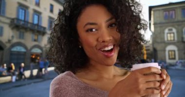 Kasaba meydanında fincandan kahve içen mutlu genç siyah kadın. Elinde içkiyle gülümseyen ve gülen güzel bir Afro-Amerikan kadını yakın çekim. 4k