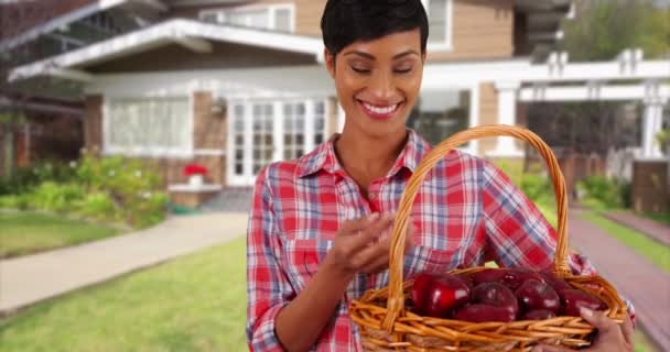陽気なアフリカ系アメリカ人女性の自家製有機果物のバスケットを保持し カメラで笑顔 美しい郊外の家の外に立って若い女性の肖像画 片手に赤いリンゴを投げ — ストック動画