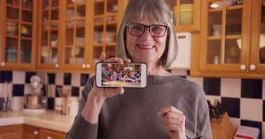 Mutlu beyaz büyükanne mutfaktaki mobil cihazda aile videosunu gösteriyor. Akıllı telefonlu neşeli, yaşlı bir bayan evin içinde kameraya aile videosu gösteriyor. 4k