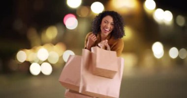 Güzel bir Afrikalı kadın alışveriş torbalarıyla bokeh ışıkları ile şehir ortamında dönüyor. Neşeli siyah kadın yeni alışverişlerle kamera şakası yapıyor. 4k