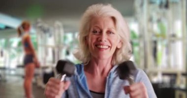 50 'li yaşlarındaki olgun bir kadın kameraya gülümseyen dambılları kaldırıyor. Spor salonunda ağırlık kaldıran güçlü yaşlı kadın. 4k