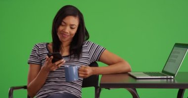 Siyah kadın, yeşil ekrandaki kafede dizüstü bilgisayarla çalışırken akıllı telefondan mesaj atıyor. Afrika kökenli Amerikalı Millennial cep telefonu uygulaması kullanıyor ve yeşil ekranda kahve ya da çay içiyor. 4k