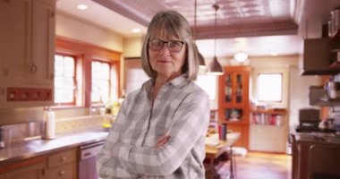 Mutfakta kolları çapraz duran kendinden emin yaşlı bir kadın. Olgun bayan temiz mutfakta kollarını kavuşturmaktan gurur duyuyor. 4k